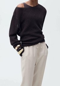 Zwarte opengewerkte trui met een oversized pasvorm en off-shoulder ontwerp, gecombineerd met lichtbeige broek met trekkoord. Soepele textuur en casual stijl.