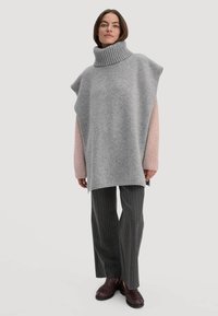 Grijze coltrui poncho met roze mouwen, oversized ontwerp, zachte textuur, gecombineerd met gestreepte grijze broek en donkerbruine instappers.