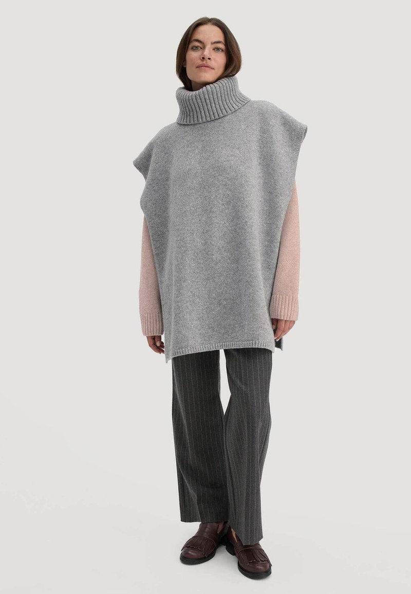 Grijze coltrui poncho met roze mouwen, oversized ontwerp, zachte textuur, gecombineerd met gestreepte grijze broek en donkerbruine instappers.