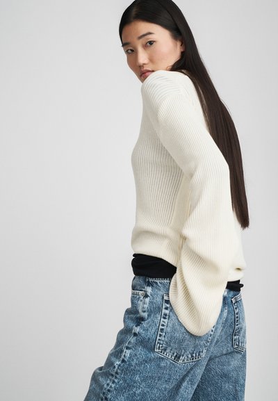 Ριγέ κρεμ πουλόβερ με μακριά μανίκια, φορεμένο πάνω από μαύρη μπλούζα, συνδυασμένο με ψηλόμεσα μπλε τζιν παντελόνια, με πίσω τσέπες.