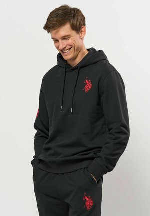 Junger Mann lächelt, trägt schwarzen Hoodie und Hose mit passenden roten Polo-Spieler-Logos, Hände in den Taschen vor einfarbigem Hintergrund.
