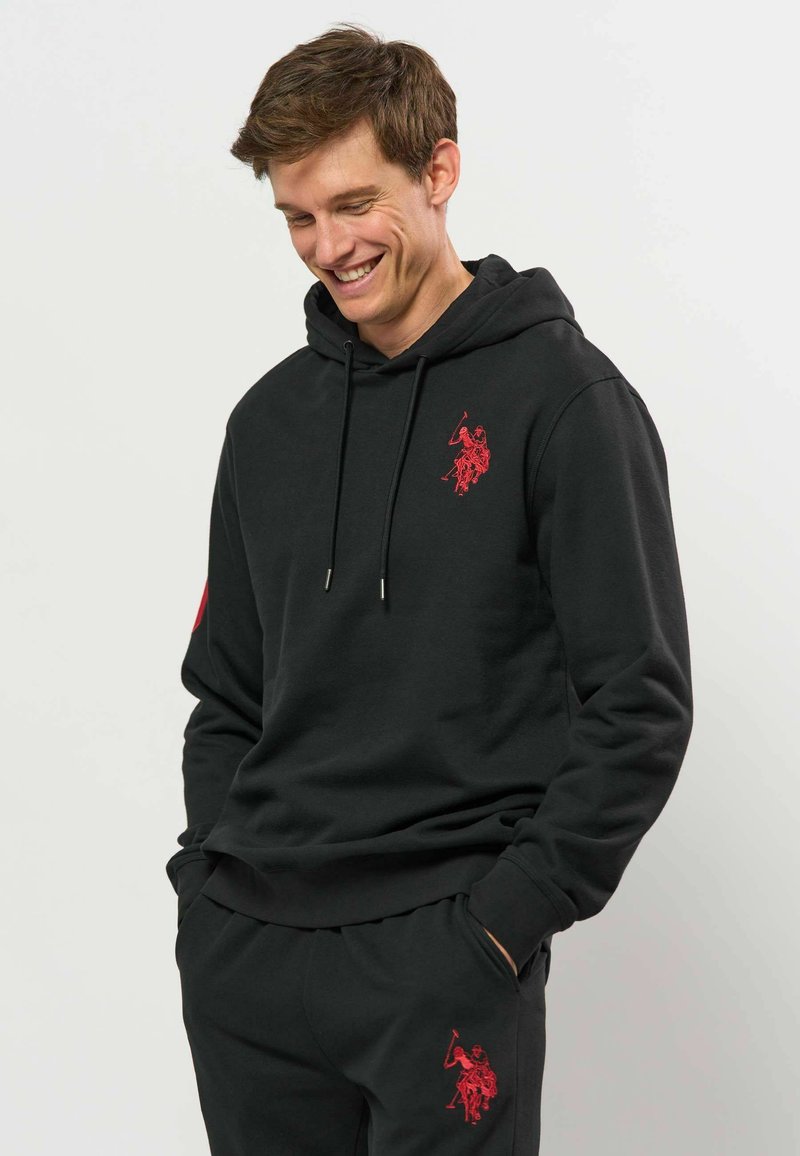 Jeune homme souriant, portant un sweat à capuche noir et un pantalon avec des logos rouges assortis de joueur de polo, mains dans les poches sur fond uni.