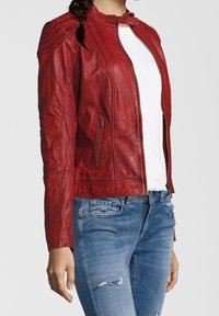 Frau in einer roten Lederjacke über einem weißen Hemd und blauen, abgetragenen Jeans, die vor einem schlichten Hintergrund steht.