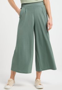 Pantalon large en tissu vert doux, avec des plis à l'avant et des poches sur les côtés. Associé à des baskets beiges. Texture lisse partout.