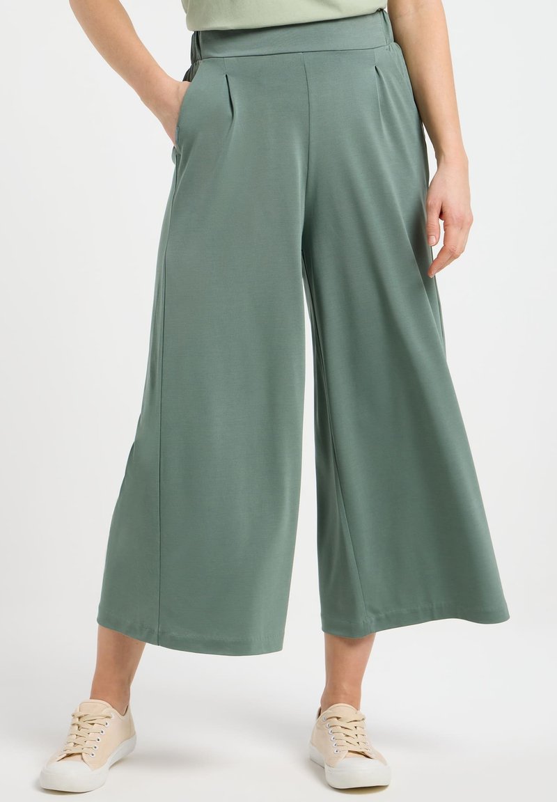 Pantalon large en tissu vert doux, avec des plis à l'avant et des poches sur les côtés. Associé à des baskets beiges. Texture lisse partout.