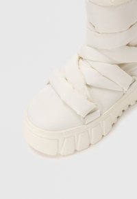 Botte blanche au design rembourré, présentant de larges sangles en tissu, un bout arrondi et une semelle épaisse et texturée. Esthétique minimaliste dans l'ensemble.