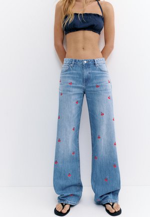 Kobieta w szerokich niebieskich dżinsach z małymi czerwonymi wzorami wiśni, czarnym topie bandeau i czarnych klapkach stoi przy białej ścianie.