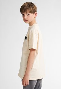Beige katoenen T-shirt met een ronde hals, korte mouwen en een zwart logopatch op de borst. De pasvorm is ontspannen en casual.