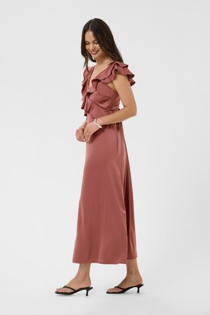 Femme en robe sans manches en satin rose poudré avec des volants, portant des sandales noires à talons, souriante et regardant vers le bas, debout sur un fond uni.
