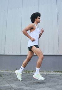 Løber i en hvid tanktop og sorte shorts, iført lysegrønne løbesko og hvide sokker, med en afro-frisure, der jogger på fortovet.