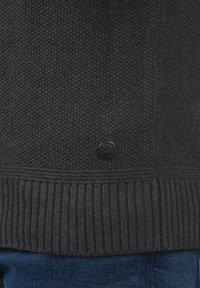 Suéter de punto gris oscuro con un patrón texturizado y dobladillo acanalado, con un botón de logo negro redondo en la parte inferior izquierda.