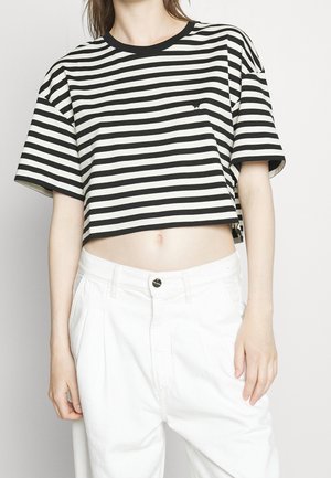 Frau trägt ein schwarz-weiß gestreiftes, kurz geschnittenes T-Shirt und hoch taillierte weiße Hose vor einem schlichten hellen Hintergrund.