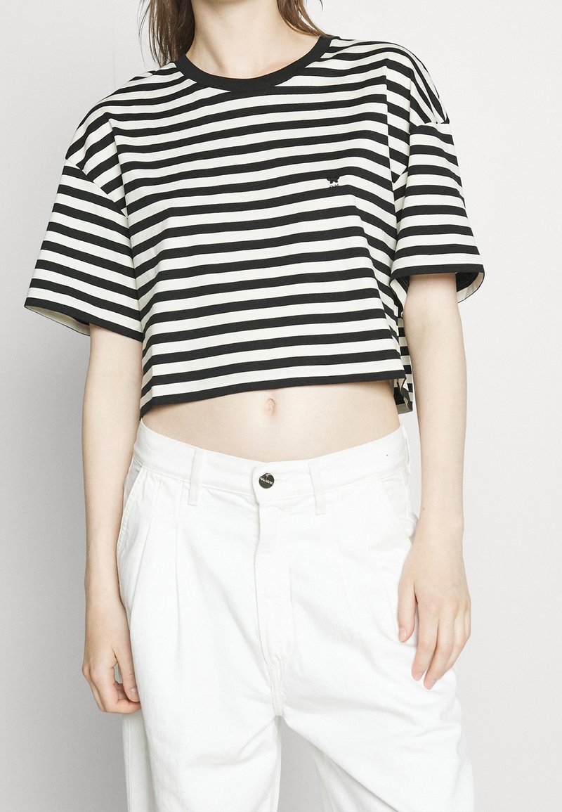 Femme portant un t-shirt court rayé noir et blanc et un pantalon blanc taille haute sur un fond clair uni.