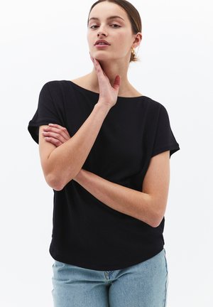 Frau mit schwarzem Kurzarm-Top und hellblauer Jeans steht mit einer Hand am Kinn und dem anderen Arm verschränkt vor einem schlichten Hintergrund.
