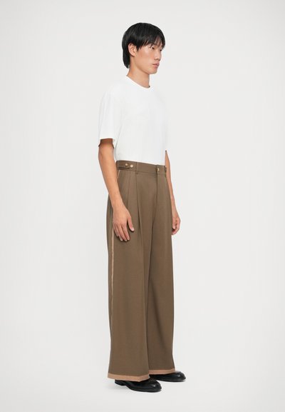Braune, weit geschnittene Hose mit hoher Taille, seitlichen Streifen und goldenen Knopf-Akzenten, kombiniert mit einem schlichten weißen T-Shirt mit Rundhalsausschnitt.