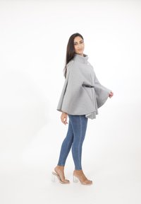 Abrigo gris con cuello alto, cierre de botones en el frente y cintura ceñida con cinturón, combinado con jeans ajustados azules y sandalias de tacón transparente.