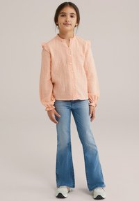 Mlada deklica stoji oblečena v breskevasto teksturirano bluzo, široke svetlo modre jeans hlače in bele superge na enotnem bež ozadju.