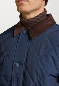 Chaquetón acolchado en color navy con un cuello de pana marrón y un cuello acanalado marrón. Presenta un cierre de botones y una textura suave y brillante.
