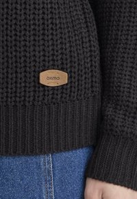 Czarny sweter z dzianiny z teksturowanym wzorem i ściągaczami, z brązową skórzaną metką z napisem "OXMO" na dolnej przedniej części.