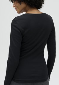 Vrouw met krullend haar, gekleed in een strakke zwarte longsleeve, zijwaarts gedraaid met gedeeltelijk zichtbaar gezicht, op een effen lichte achtergrond.