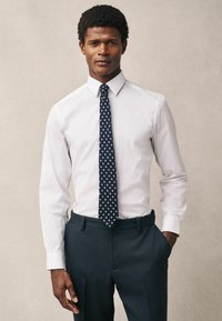 Uomo che indossa una camicia bianca, cravatta blu navy con pois bianchi e pantaloni scuri, in piedi con una mano in tasca su uno sfondo neutro.