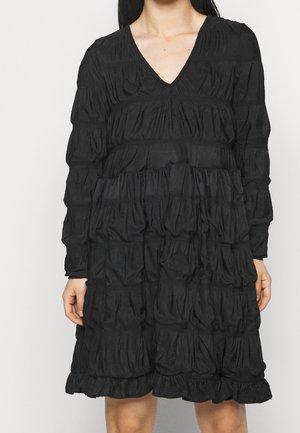Femme portant une robe noire à manches longues avec un col en V et une texture de tissu froncée en étages.