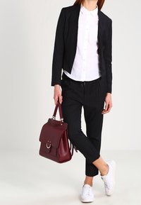 Žena v černém pruhovaném obleku a bílé košili, držící vínovou kabelku, stojící s jednou nohou překříženou, oblečená v bílých teniskách.