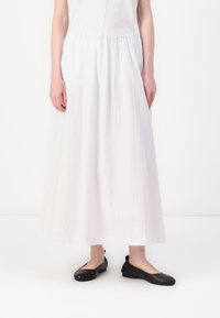 Jupe blanche longueur cheville en tissu léger avec une taille élastique ; assortie à des chaussures plates noires. Design simple, sans motifs ni accents.