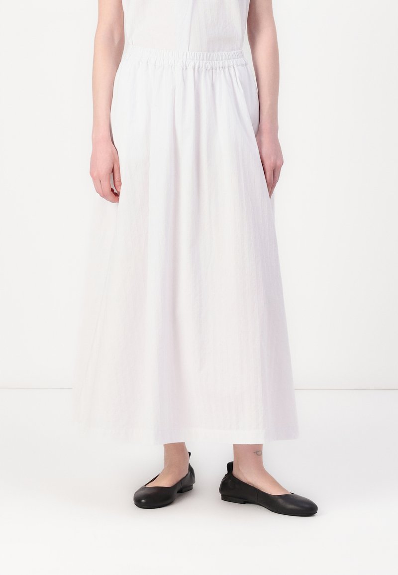 Jupe blanche longueur cheville en tissu léger avec une taille élastique ; assortie à des chaussures plates noires. Design simple, sans motifs ni accents.