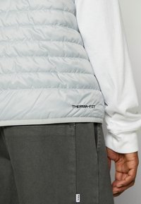 Ljusgrå pufferväst med horisontell quiltad design, som har en svart "THERMA-FIT"-logotyp och är kombinerad med mörkgrå byxor av texturerad tyg.