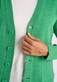 Groene cardigan gemaakt van gebreid materiaal, met geribbelde randen en grote turquoise knopen. Hand geplaatst bij het knoopsgat aan de rechterkant.