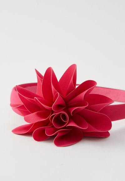 Un bandeau en tissu rose avec un grand accent floral, comprenant des pétales superposés et une finition texturée, maintenu par un bandeau doux et extensible.