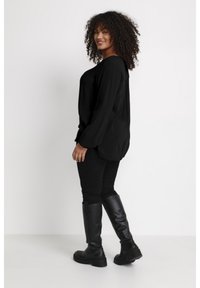 Haut noir à manches longues avec un ourlet arrondi, associé à un jean skinny noir et des bottes noires brillantes montant jusqu'aux genoux. Cheveux bouclés et longueur aux épaules.