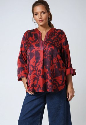 Vrouw draagt een losse rood-zwarte blouse met bloemenprint, opgerolde mouwen en wijde blauwe spijkerbroek tegen een effen achtergrond.