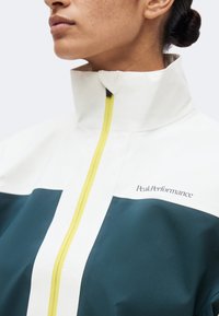 Άτομο που φοράει μπουφάν με ψηλό γιακά, κίτρινο φερμουάρ, λευκή πάνω περιοχή, σκουρόχρωμη πετρόλ κάτω περιοχή και το λογότυπο "Peak Performance" στο στήθος.