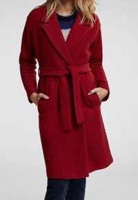 Manteau rouge ceinturé avec manches longues, revers crantés et poches latérales. Tissu texturé avec une coupe légèrement ample, arrivant au-dessus du genou.