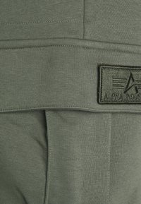 Olivgrüner Baumwollmischung-Hoodie mit geripptem Muster, ausgestattet mit einem schwarzen gestickten Etikett mit "ALPHA INDUSTRIES" auf der Vorderseite.