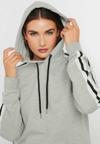 Sudadera gris con capucha y cordones negros, con rayas en blanco y negro en las mangas. Textura suave y corte relajado.