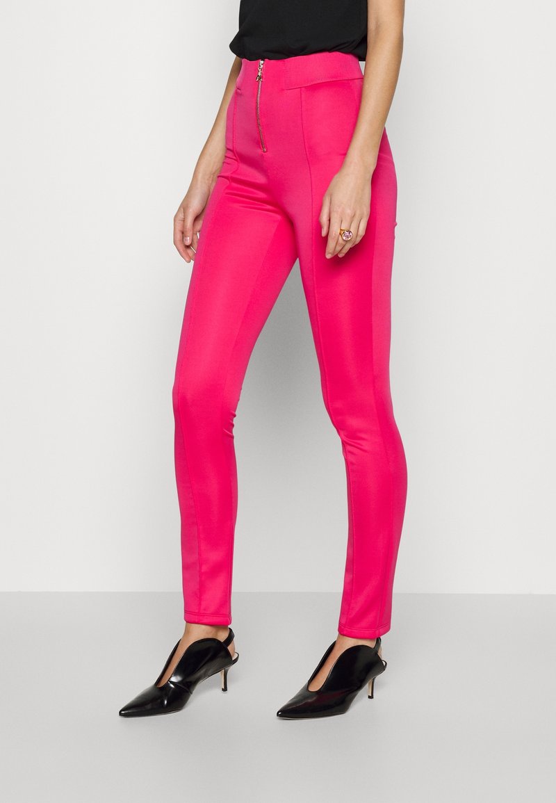 Femme portant un pantalon slim taille haute rose vif avec fermeture éclair à l'avant, un haut noir sans manches, des escarpins noirs à bout pointu avec bride arrière, et une bague.