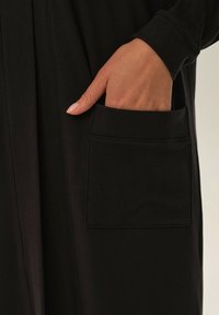 Czarna odzież z boczną kieszenią, charakteryzująca się gładką teksturą i dopasowanym mankietem. Widoczna ręka spoczywająca w kieszeni dla porównania wielkości.