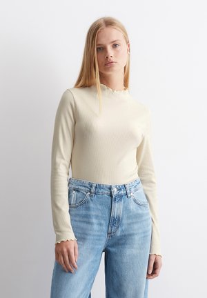 Haut à manches longues en ribbed beige avec un col festonné. Porté avec un jean bleu clair taille haute, présentant une coupe décontractée.