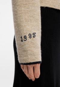 Πλεκτό μπεζ πουλόβερ με μανίκια που διαθέτουν μαύρες λεπτομέρειες και το έτος "1985" κεντημένο μαύρα στη μανσέτα. Υφή, ζεστό και εφαρμοστό σχέδιο.