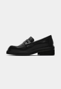 Mocassin en cuir noir avec un petit talon bloc, bout arrondi, et un détail de lanière sur le dessus.