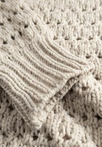 Tekstuuriga, paksust lõngast pleed pehmes beežis varjus, millel on soonikserv ja palmikumustrid, mis näitavad sooja ja kutsuvat kangast.