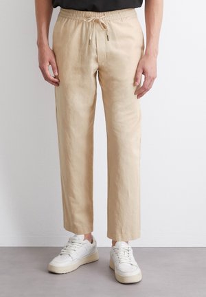 Pantalon beige droit avec taille élastique et cordon, porté avec des baskets blanches par une personne debout devant un mur uni.
