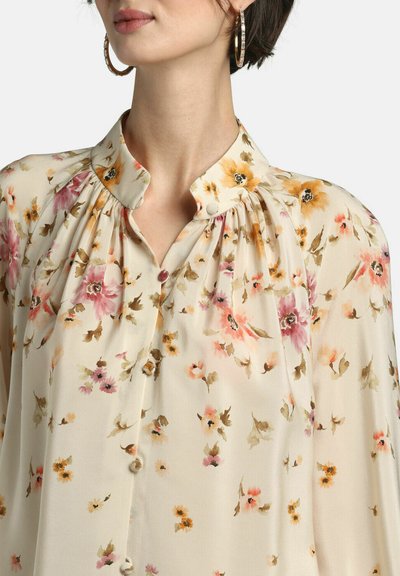 Woman wearing a cream blouse with yellow, pink, and orange floral print and a mandarin collar, paired with large gold hoop earrings.