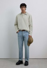 Camisa polo de manga larga color beige claro, jeans azul claros, zapatos negros, sosteniendo una gorra marrón. Diseño sencillo con colores neutros y corte clásico.
