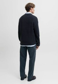 Dunkelgrauer Strickpullover mit strukturierter Oberfläche, über einem hellblauen Hemd gezogen, kombiniert mit dunkelgrünen Hosen und schwarzen Schuhen.