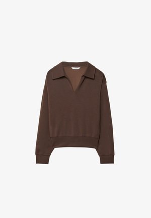 Persona con pantalones anchos de color marrón oscuro, una parte superior marrón oscuro a juego con cuello, sosteniendo un bolso marrón, de pie en un suelo gris.