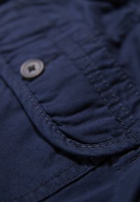 Navyblått tyg i närbild, med en texturerad yta, framträdande ficka och en silverfärgad knapp med ett korsdesign.